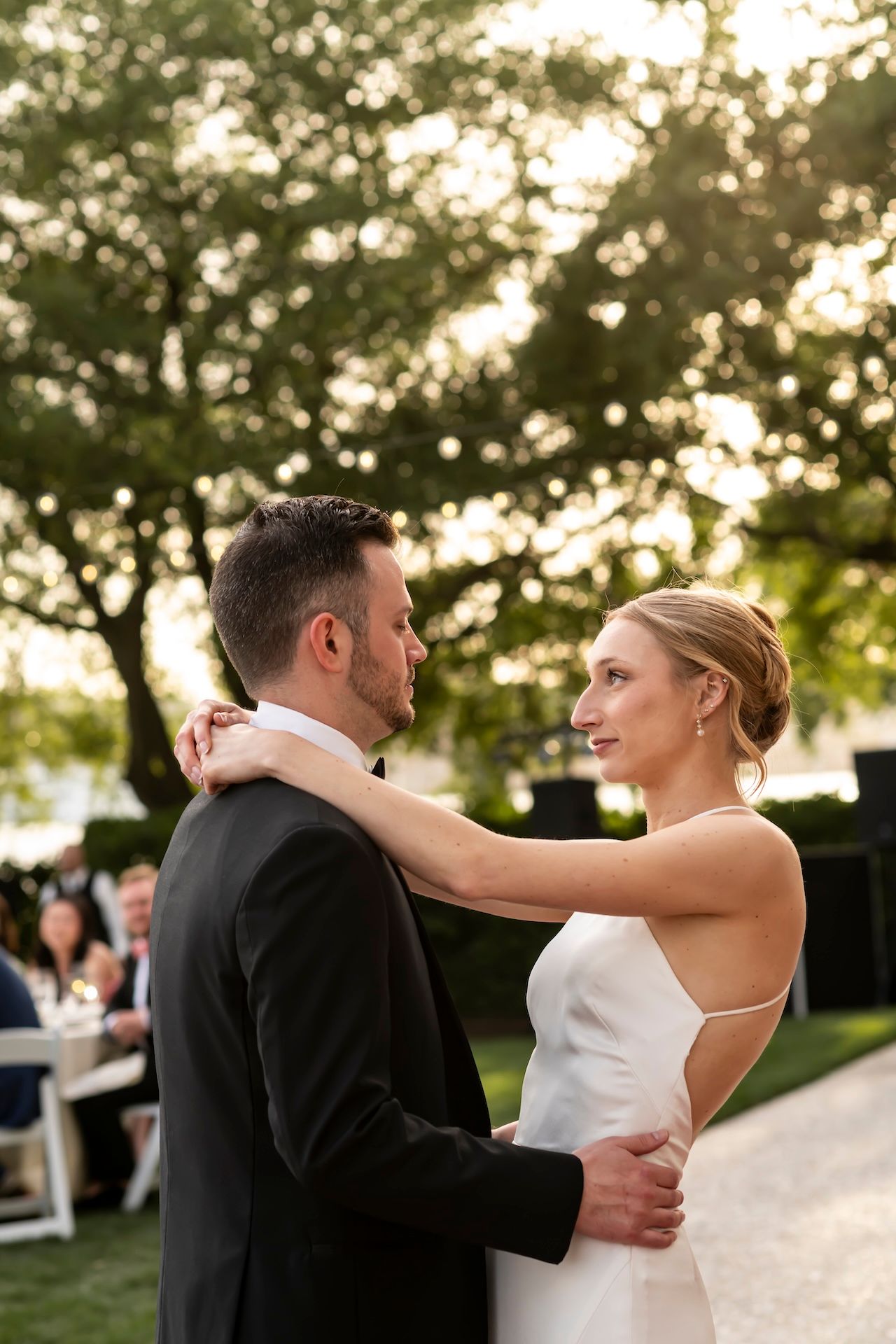 Bride and groom share their first dance outdoors at golden hour, surrounded by lush trees and wedding guests.