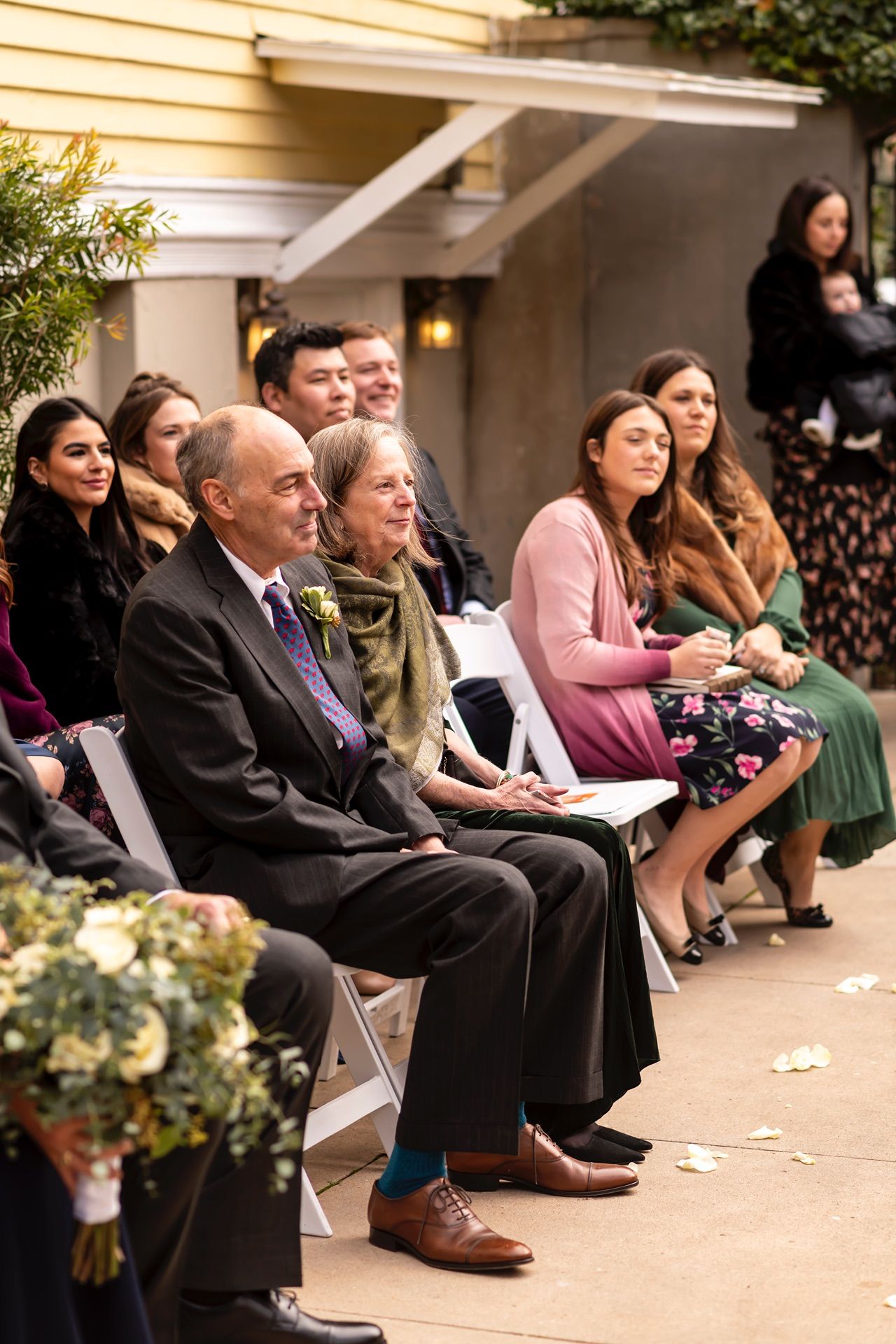 Row of attendees seated at an outdoor event watching and listening attentively in a covered patio area.