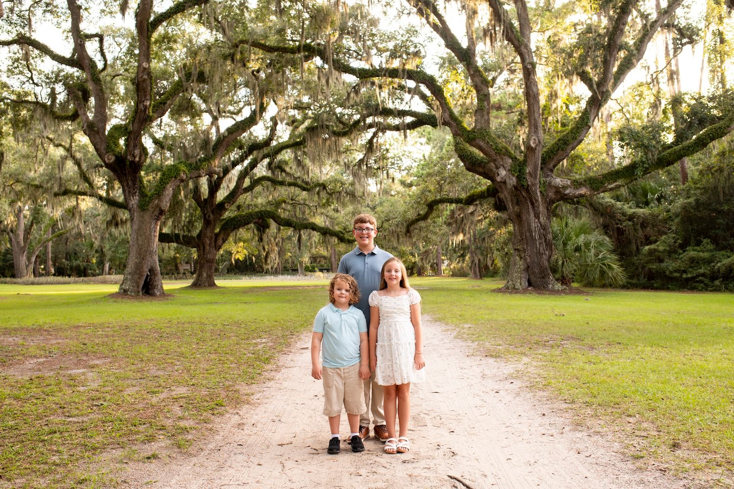 A father poses with his two young children on a scenic tree-lined path outdoors.