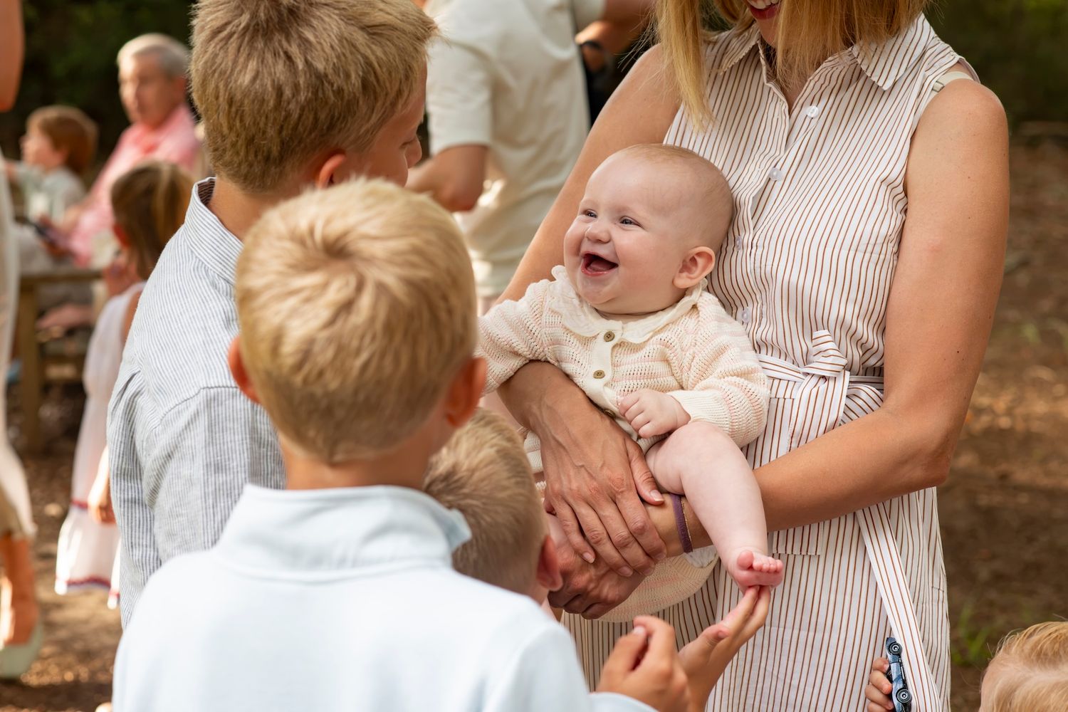 A laughing baby being held by a woman in a striped dress surrounded by children at an outdoor family gathering.