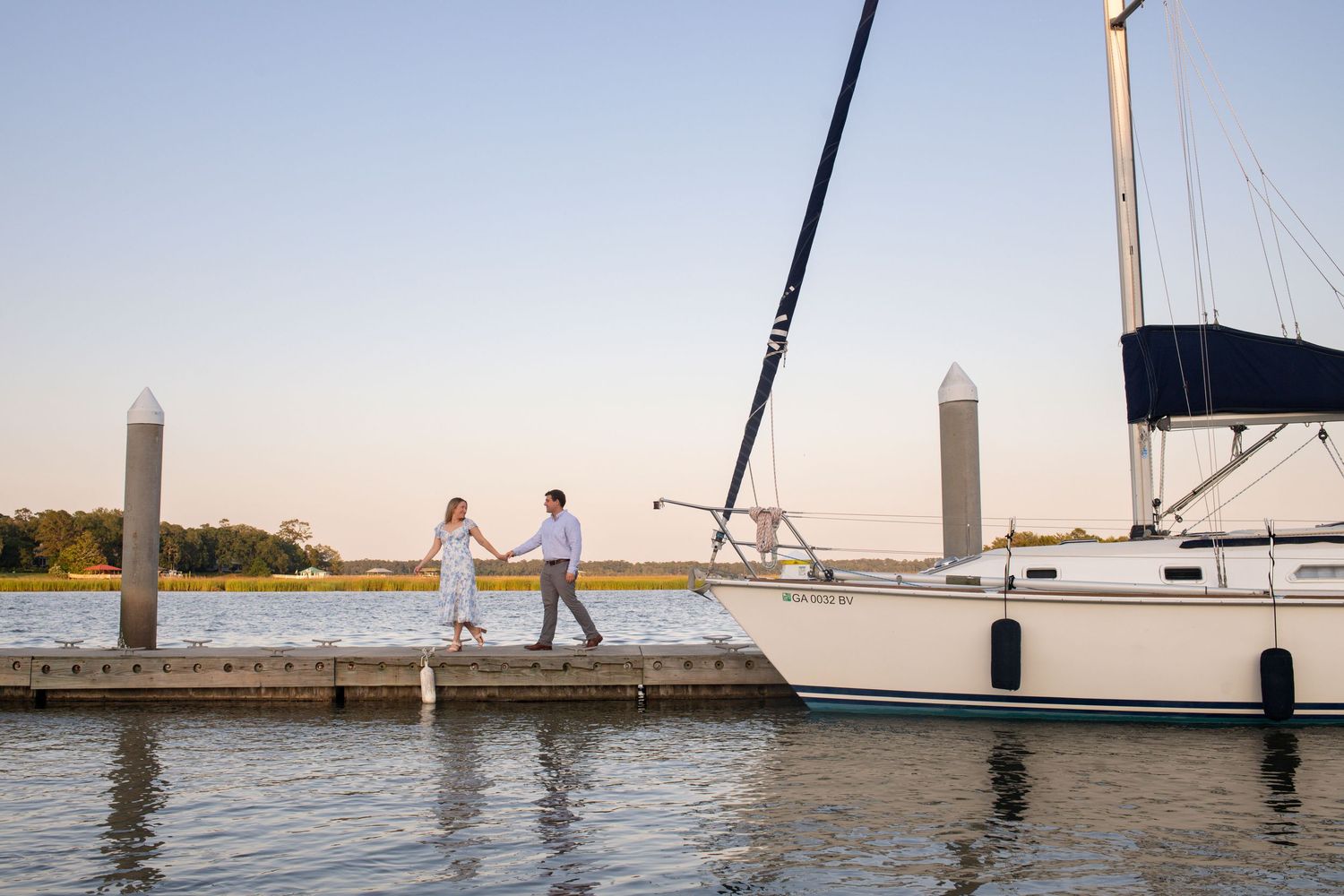 Couple walking on pier at sunset near docked sailboats at marina.