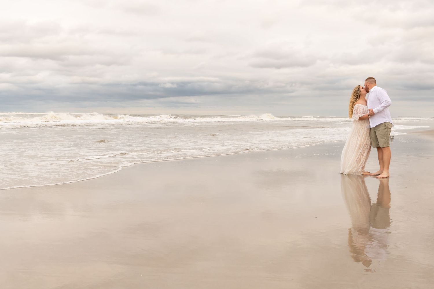 A couple embraces on a serene beach with waves and cloudy skies in the background.