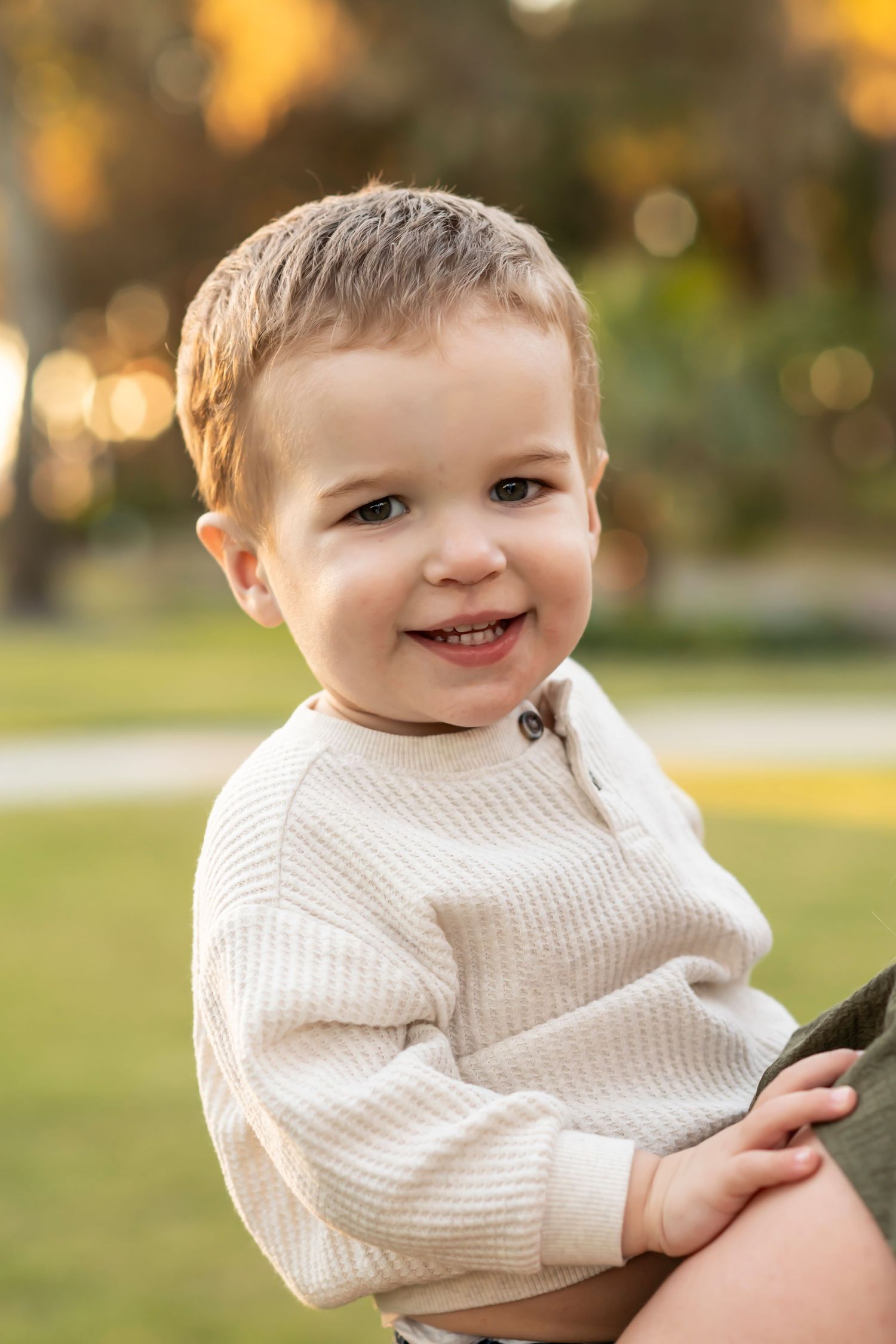 Young child in cream sweater smiles brightly during outdoor golden hour photo session.