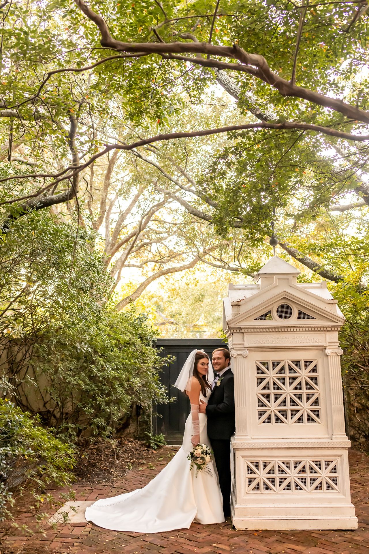 A romantic couple embraces on a garden path with white decorative stone architectural elements and lush greenery.