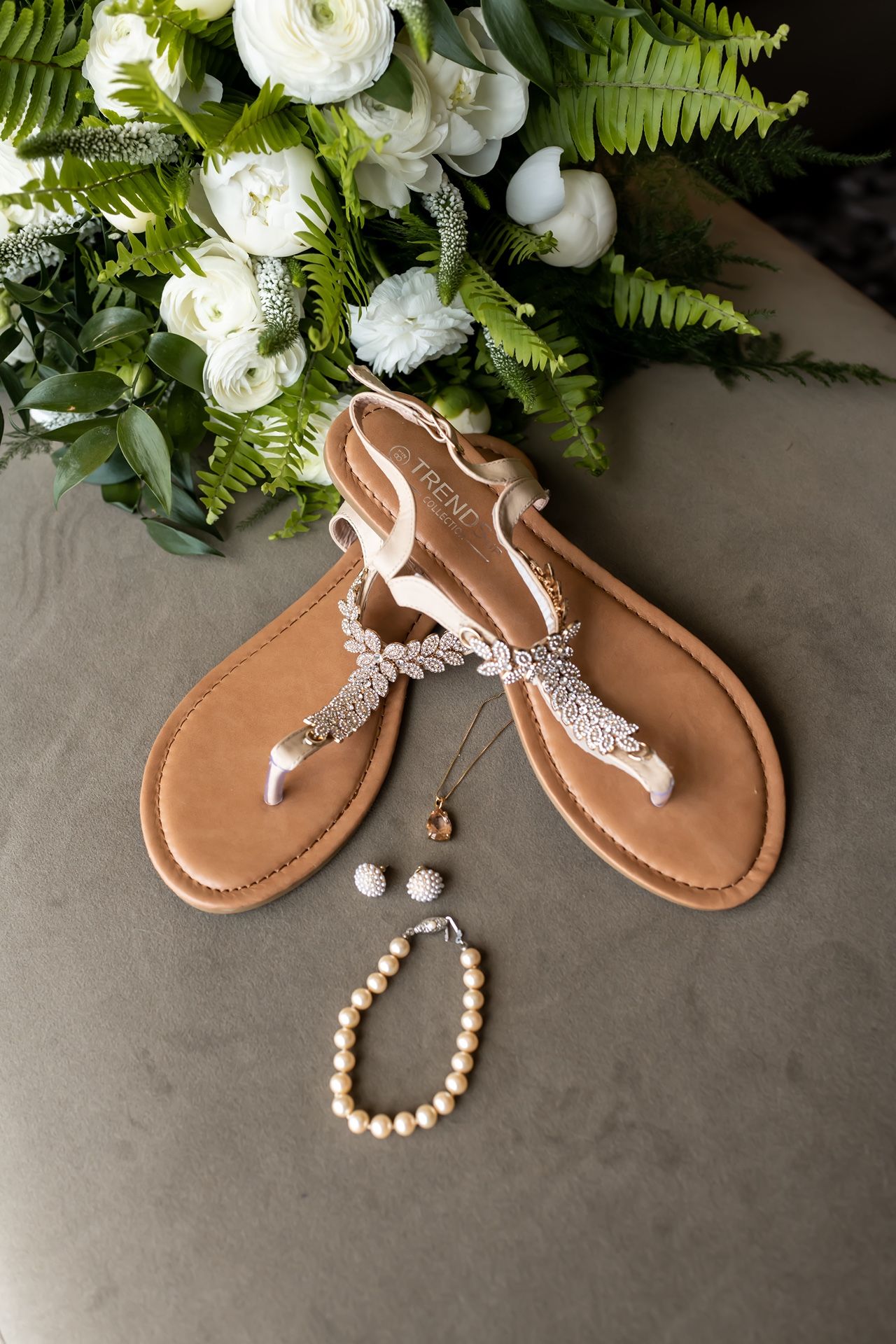 Wedding accessories including sparkly sandals, pearl bracelet and jewelry arranged with white flowers and green ferns.