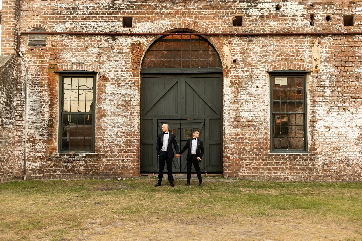 Two people in formal attire stand in front of a weathered brick building with large dark green arched door and windows.