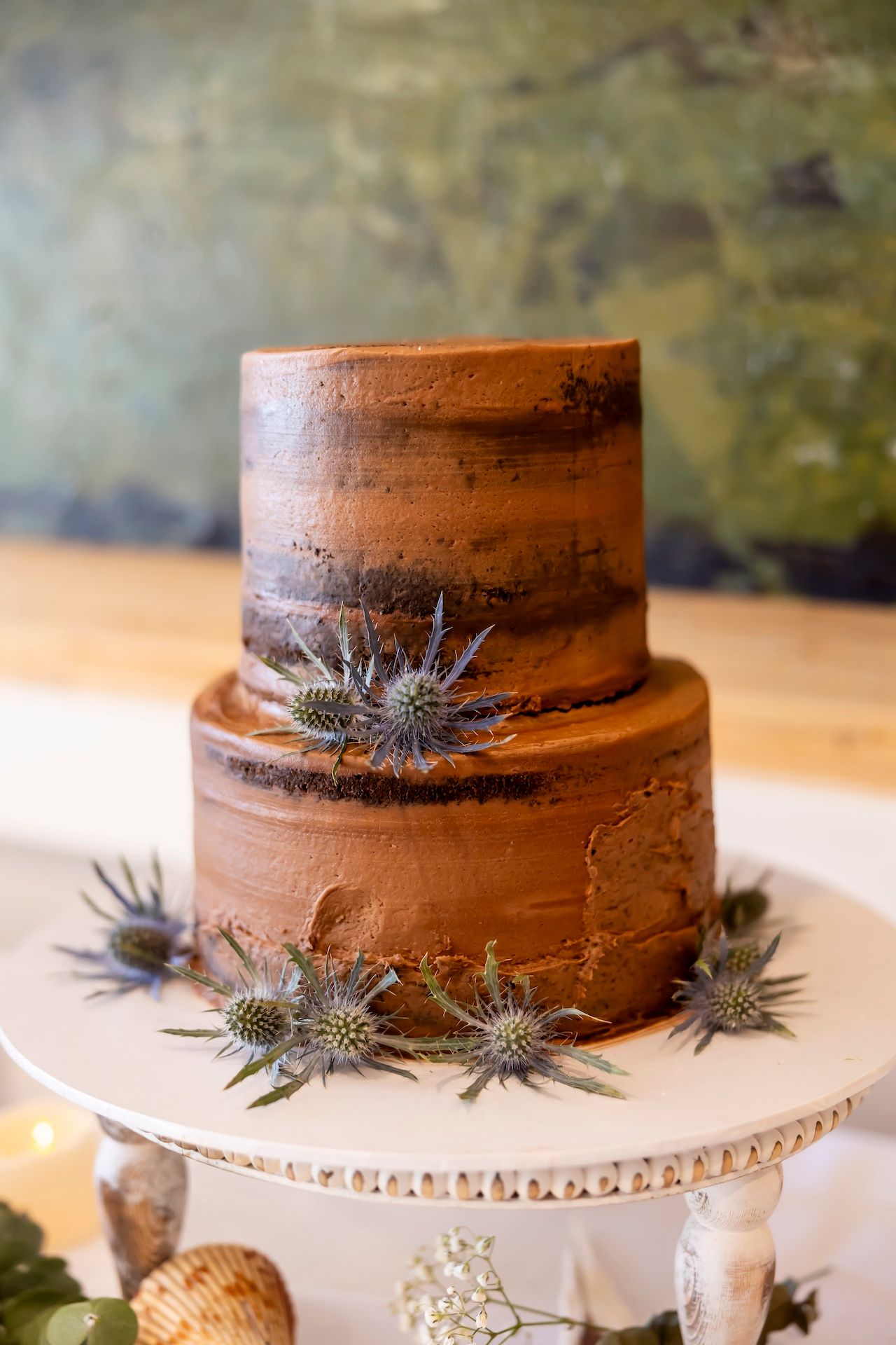 Two-tiered chocolate wedding cake decorated with spiky thistle flowers on a white pedestal cake stand.