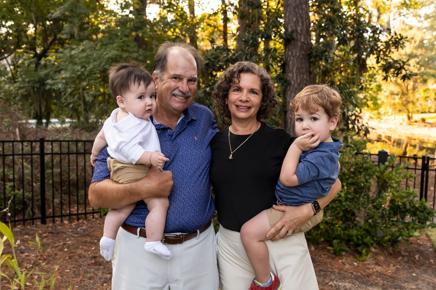 A family of four poses together outdoors at sunset in front of trees and a fence.