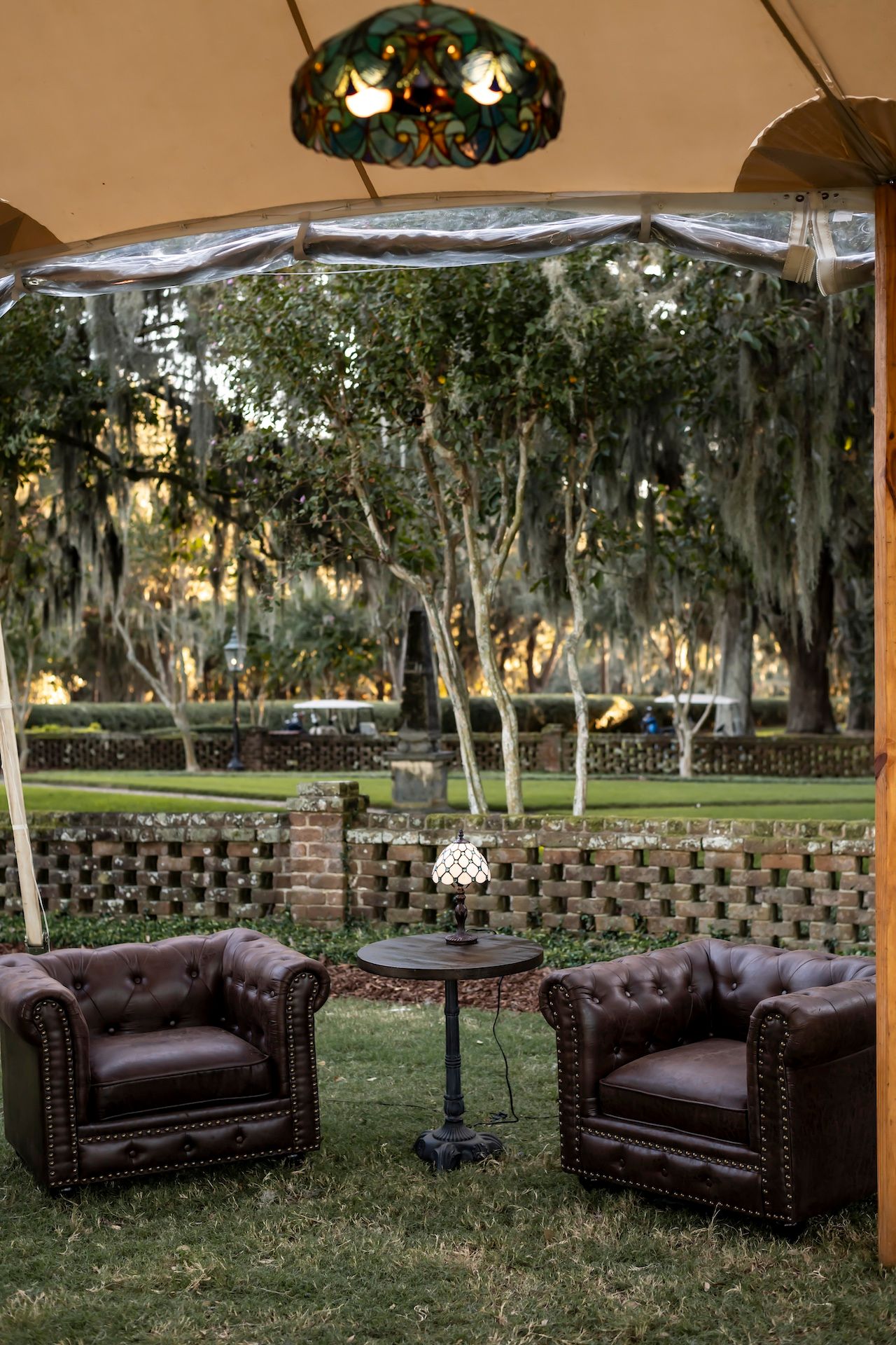 Elegant leather tufted armchairs and side table set up outdoors under a white canopy with Spanish moss draped trees in background.