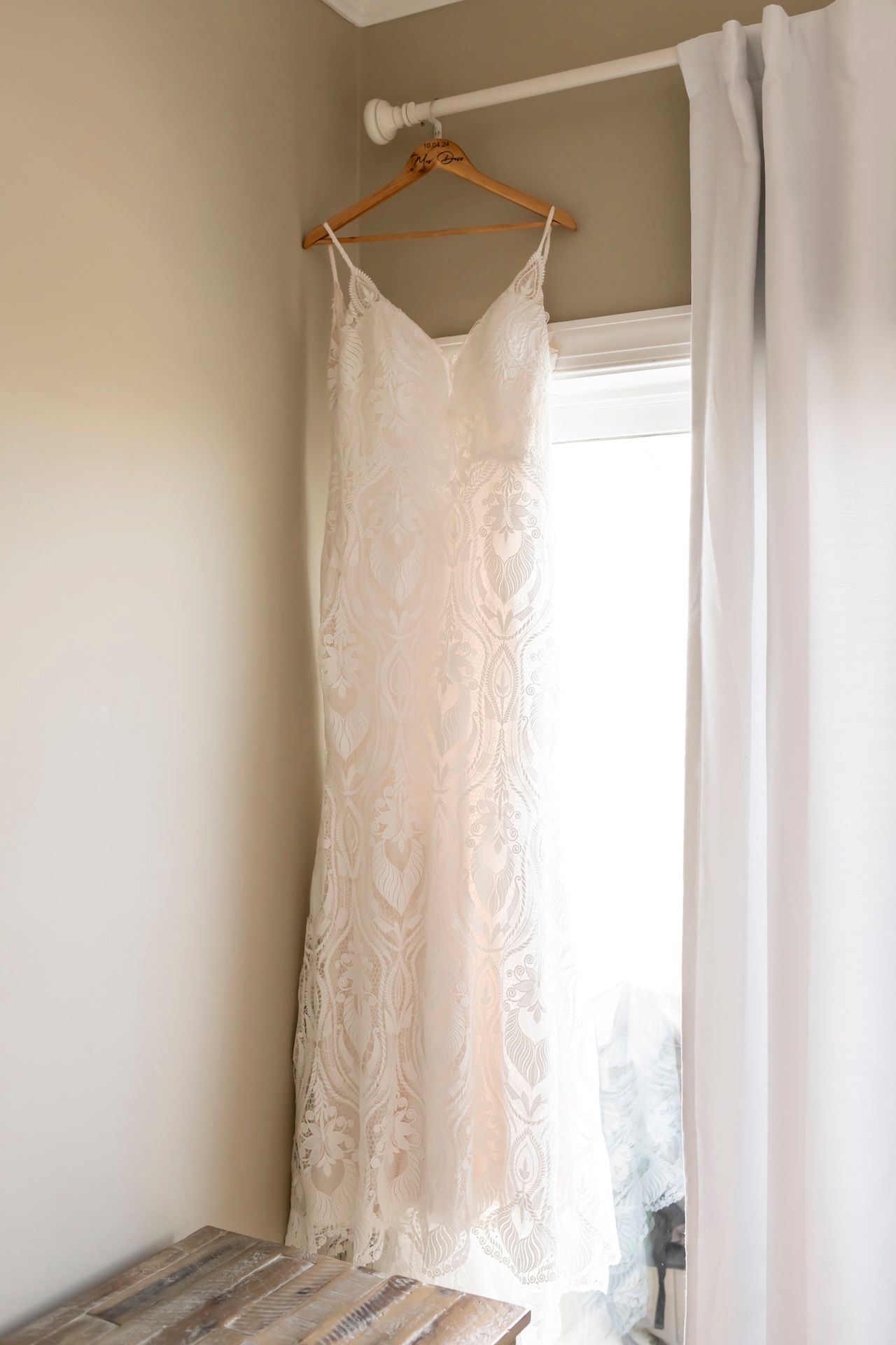 An elegant white lace wedding dress with spaghetti straps hangs on a wooden hanger by a sunlit window.