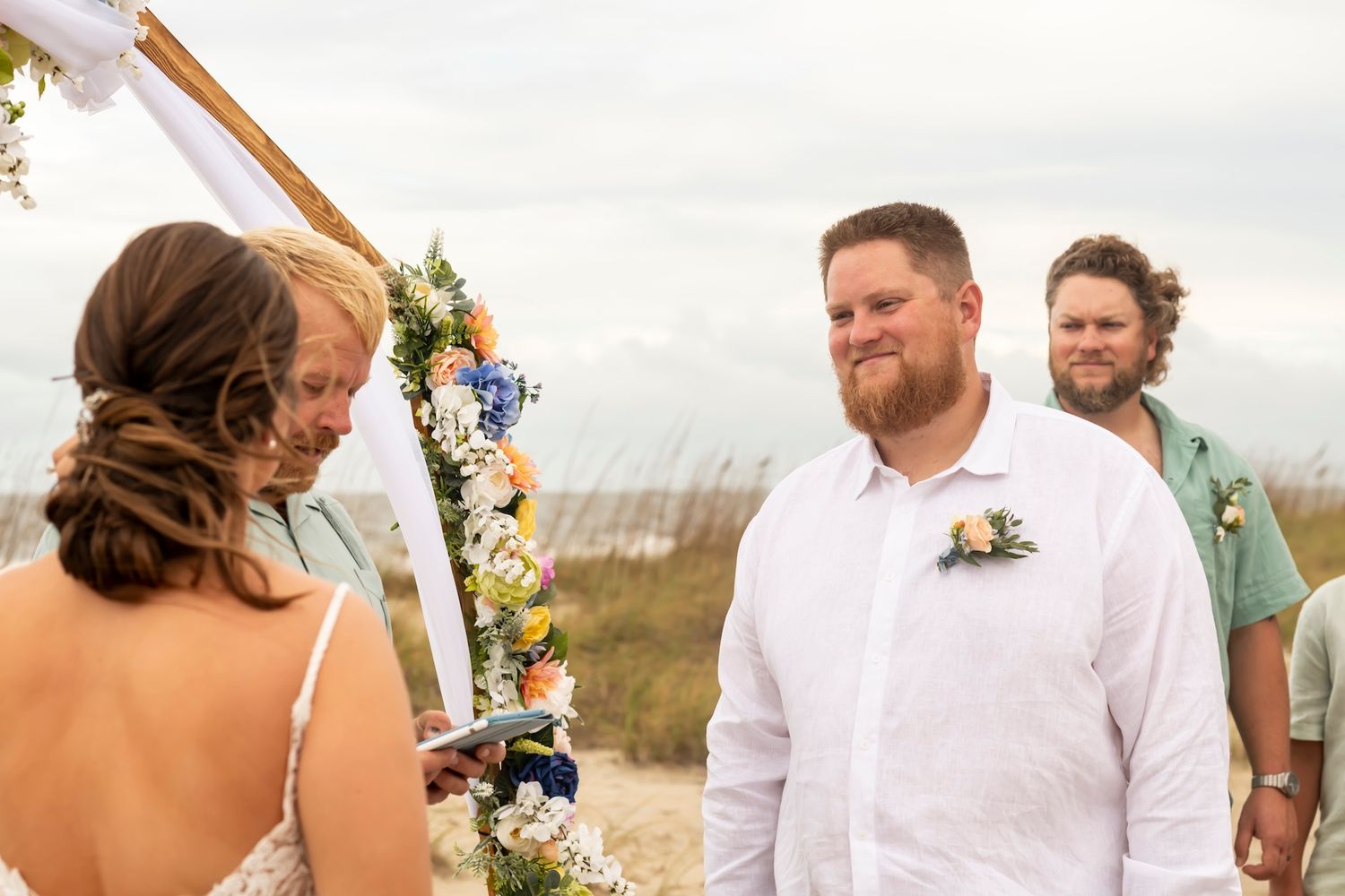 An outdoor beach wedding ceremony features a decorated wooden arch and floral decorations against a coastal backdrop.
