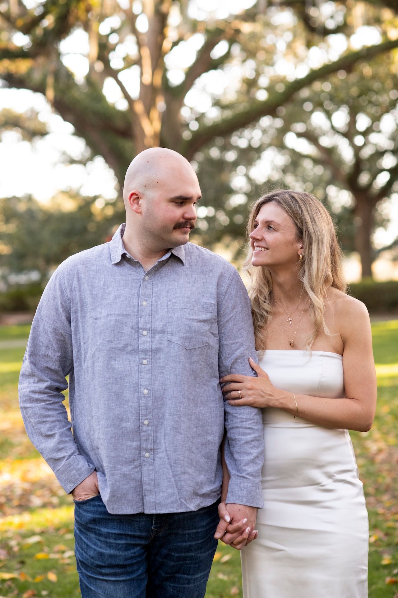 A couple holds hands and gazes at each other lovingly in a park setting with golden sunlight filtering through trees.