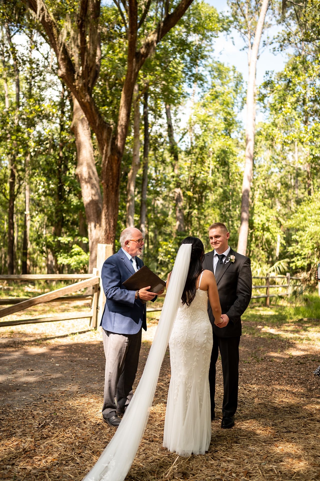 An outdoor wedding ceremony takes place in a sunlit wooded area with tall trees and natural ground cover.