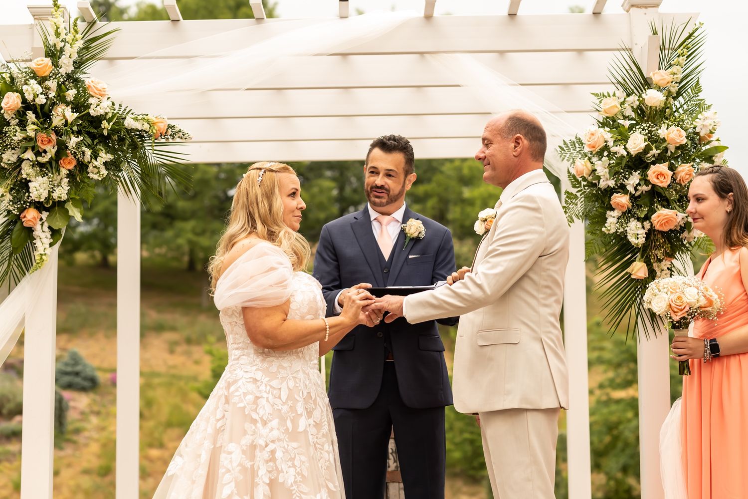 Wedding ceremony takes place under white pergola adorned with peach and cream floral arrangements.