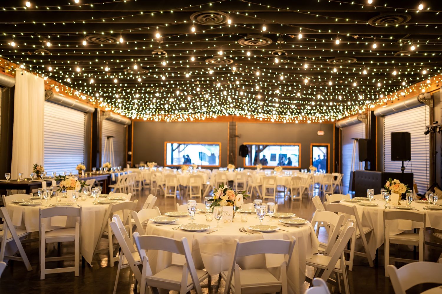 An elegant wedding reception venue with twinkling fairy lights overhead and round tables with white linens and chairs.