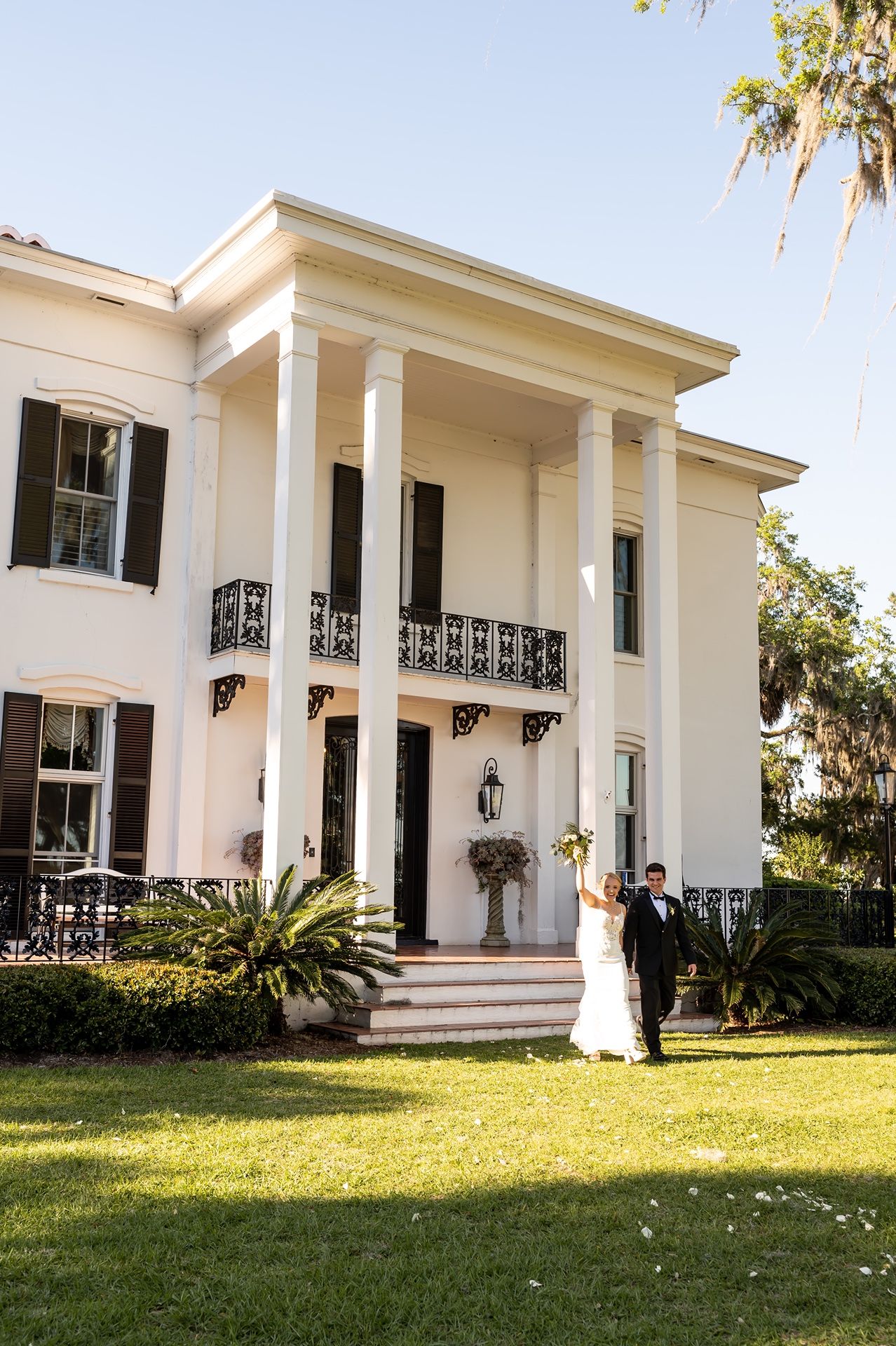 Historic Southern mansion with white columns and ornate iron railings features a wedding couple standing on the front steps.
