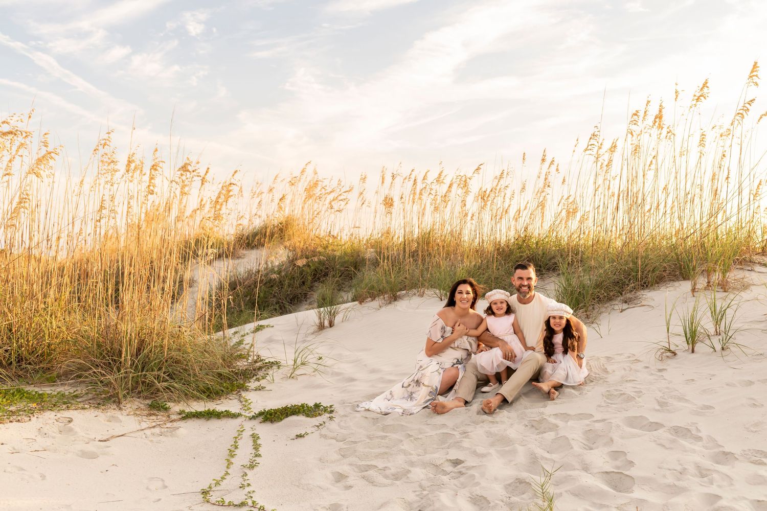Family relaxing together on sandy beach dunes with tall sea grass swaying in golden sunset light at shore.