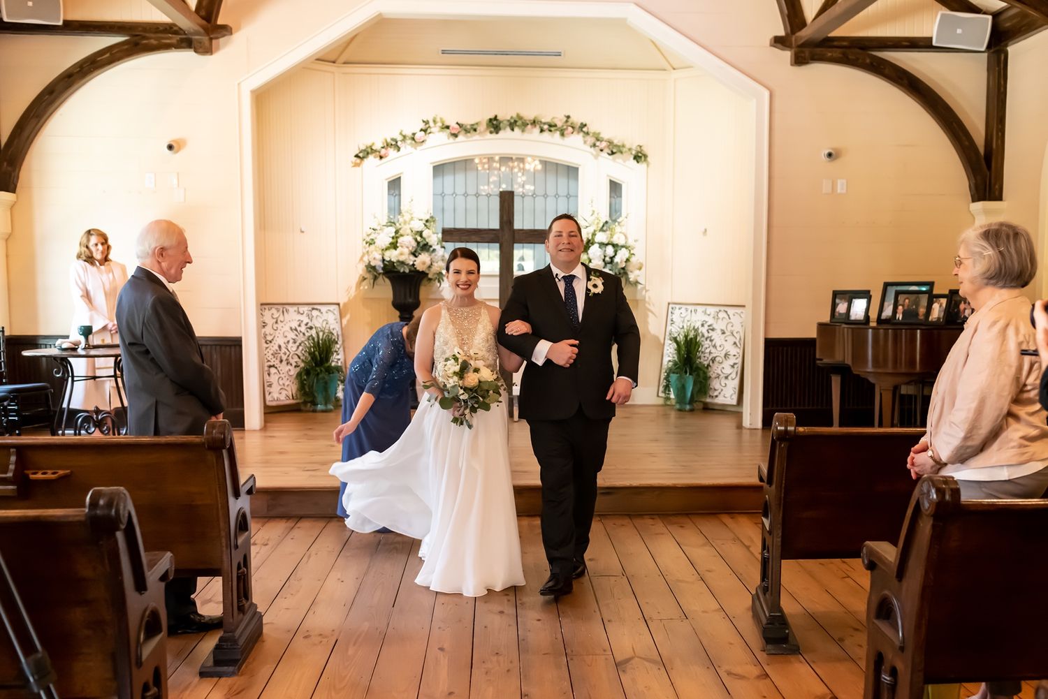 Couple walks down wooden aisle together in rustic wedding chapel with decorative floral arch and wooden pews.