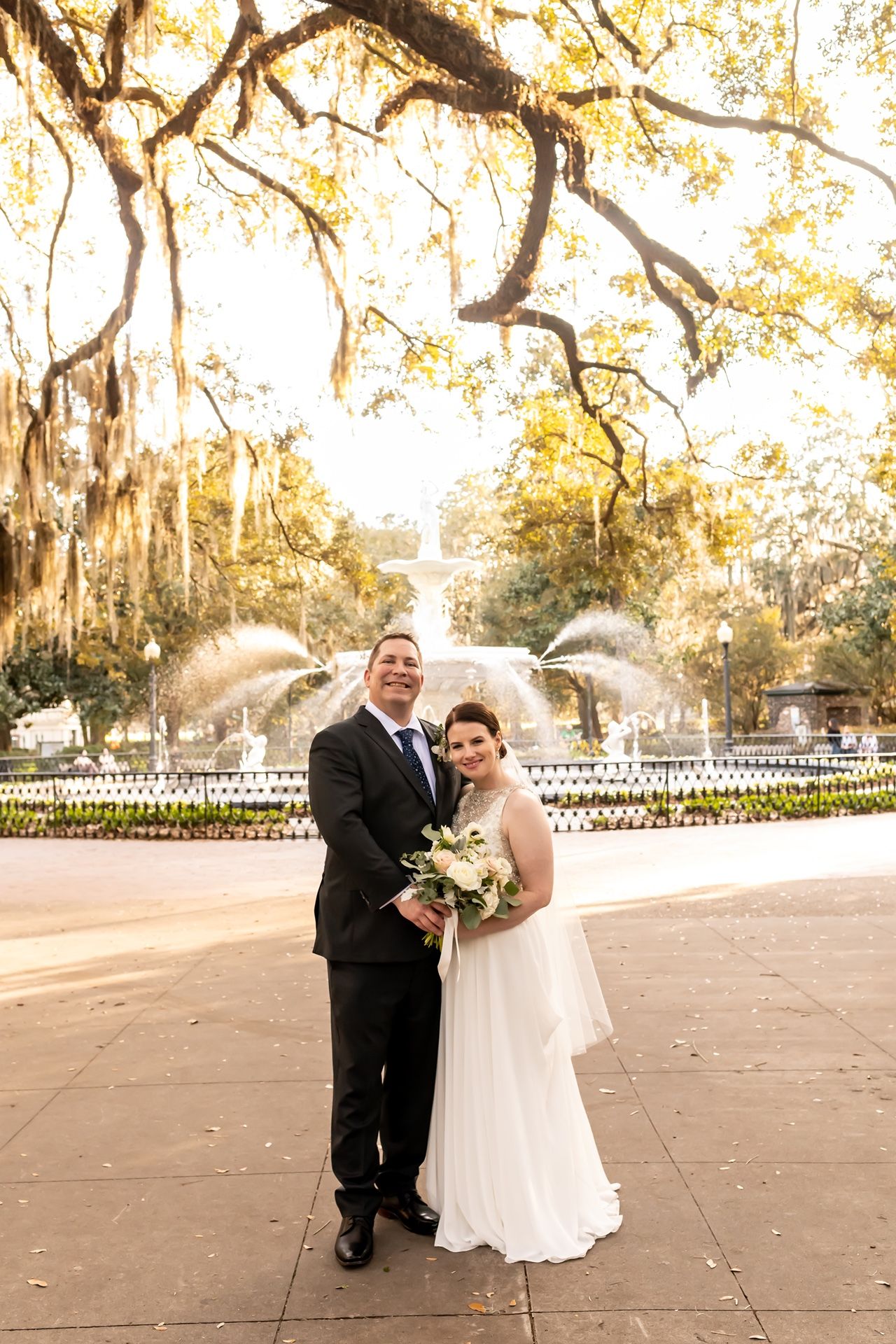 A bride and groom pose for wedding photos beneath Spanish moss-draped oak trees in a historic park.
