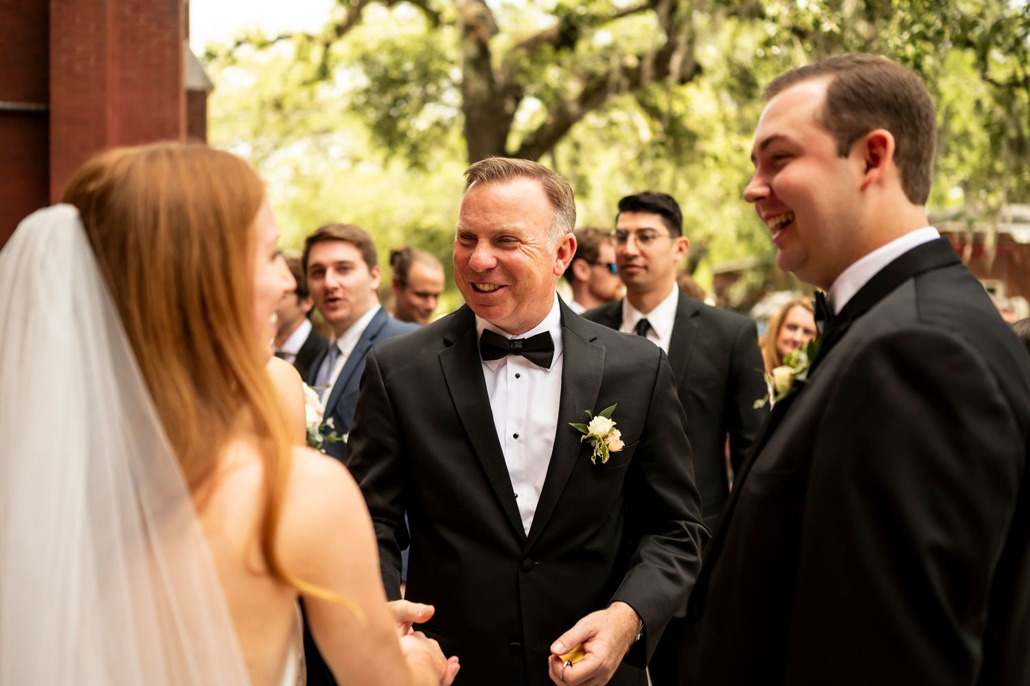 Wedding ceremony attendees share a joyful moment outdoors with sunshine filtering through trees in the background.