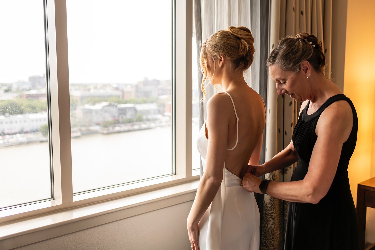 A person in black helps adjust the back of an elegant white dress near a large window with a cityscape view.