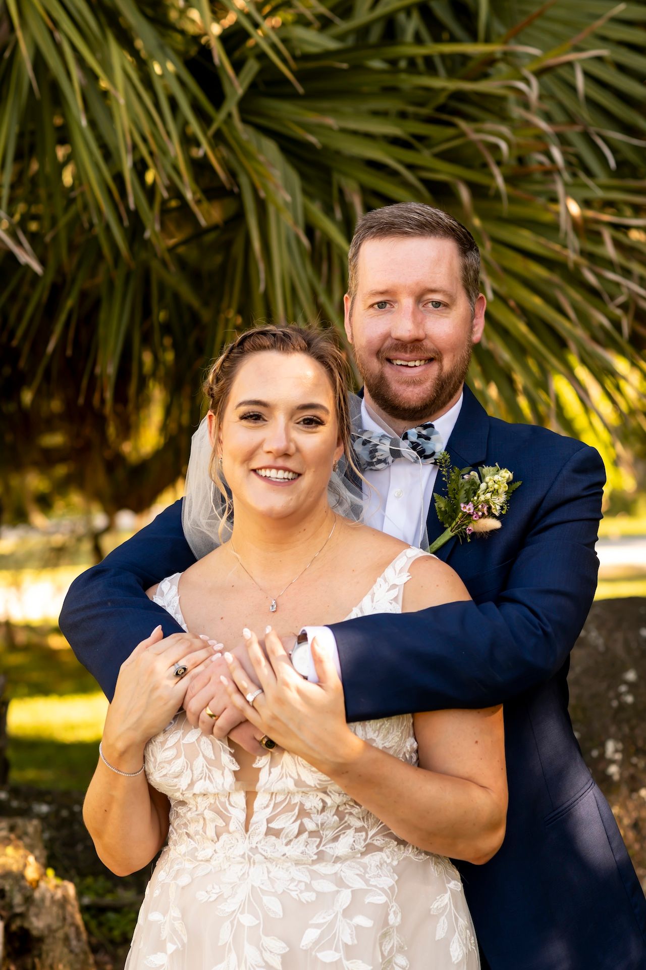 A romantic outdoor wedding portrait with the couple embracing, featuring lush palm trees in the background.
