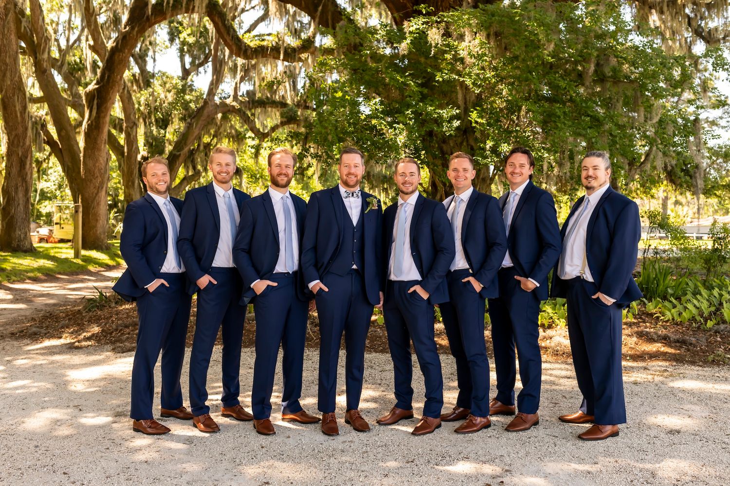Groomsmen in navy suits and brown shoes pose for creative group photos outdoors under sunlit trees.