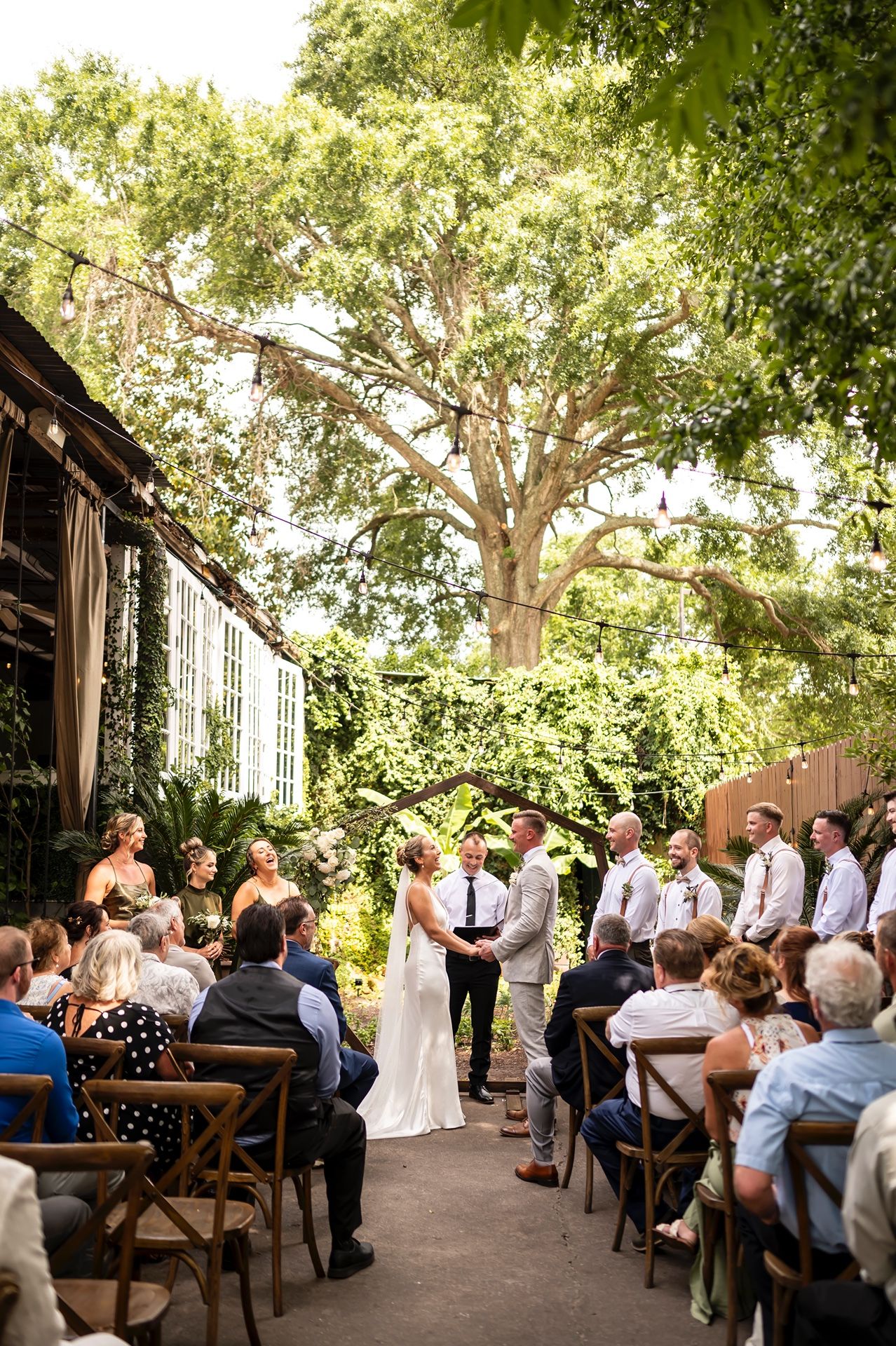 Intimate outdoor garden wedding ceremony set under a large shady tree with rustic wooden chairs arranged on a patio.