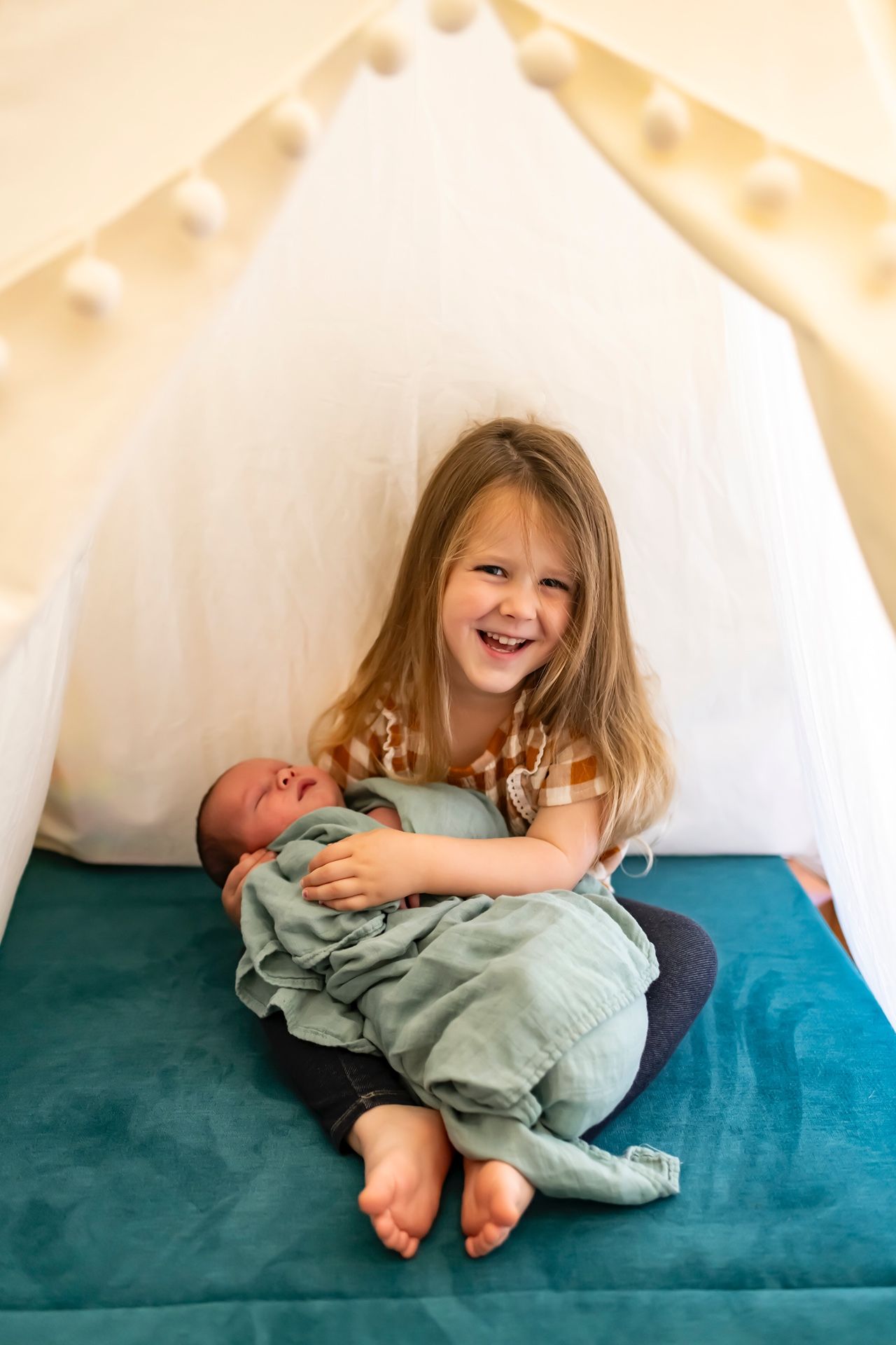 Two young children sit together inside a cozy tent with string lights, one holding a baby on a teal cushion.