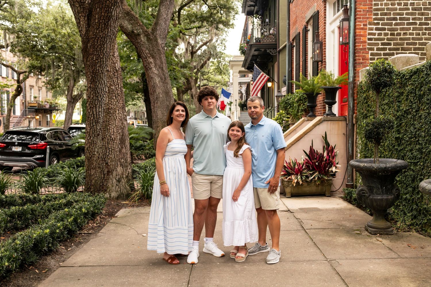 A group in white summer outfits stand together on a historic brick-lined street with oak trees overhead in Savannah.