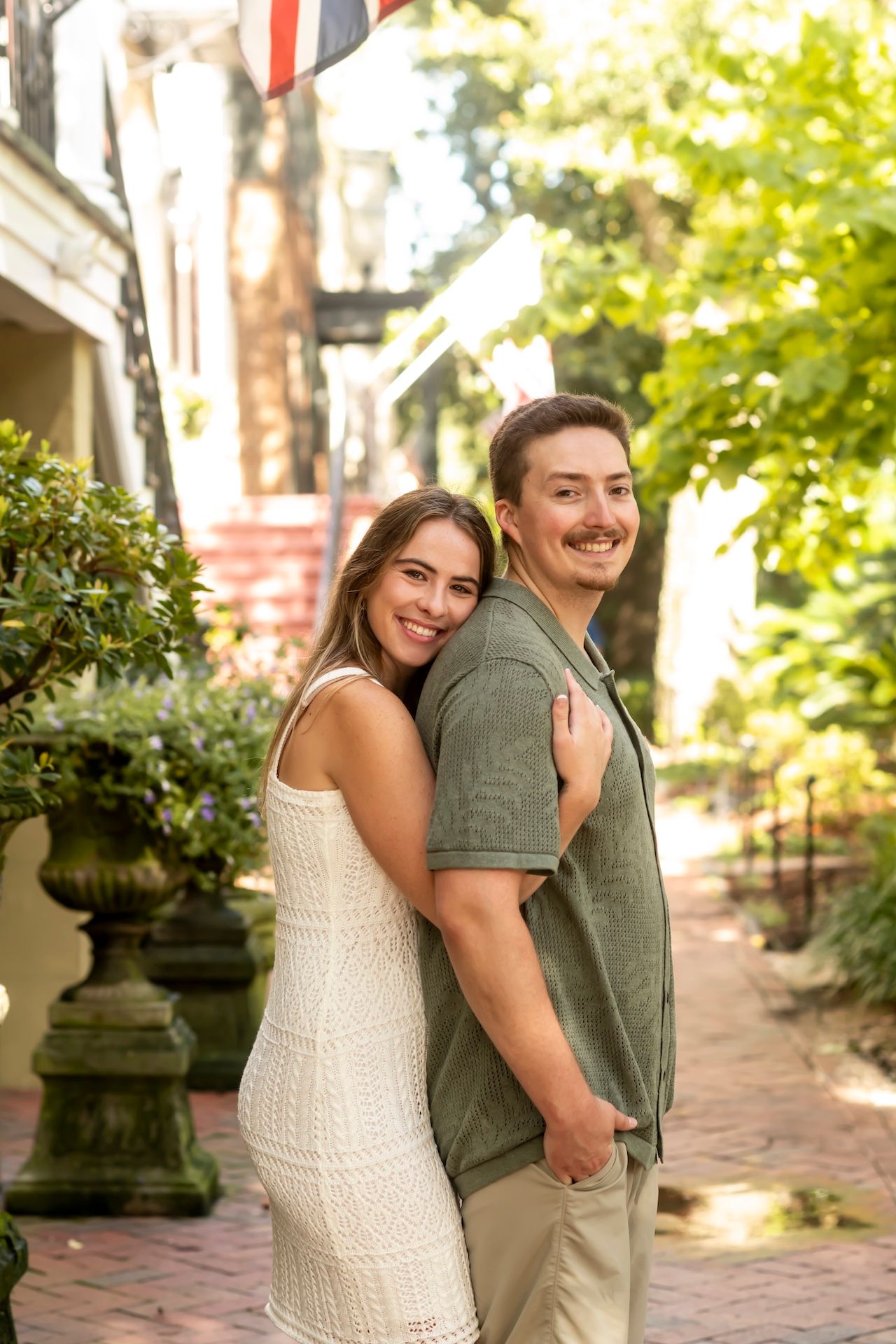 A couple embraces and laughs together while standing on a brick pathway surrounded by greenery and garden urns.