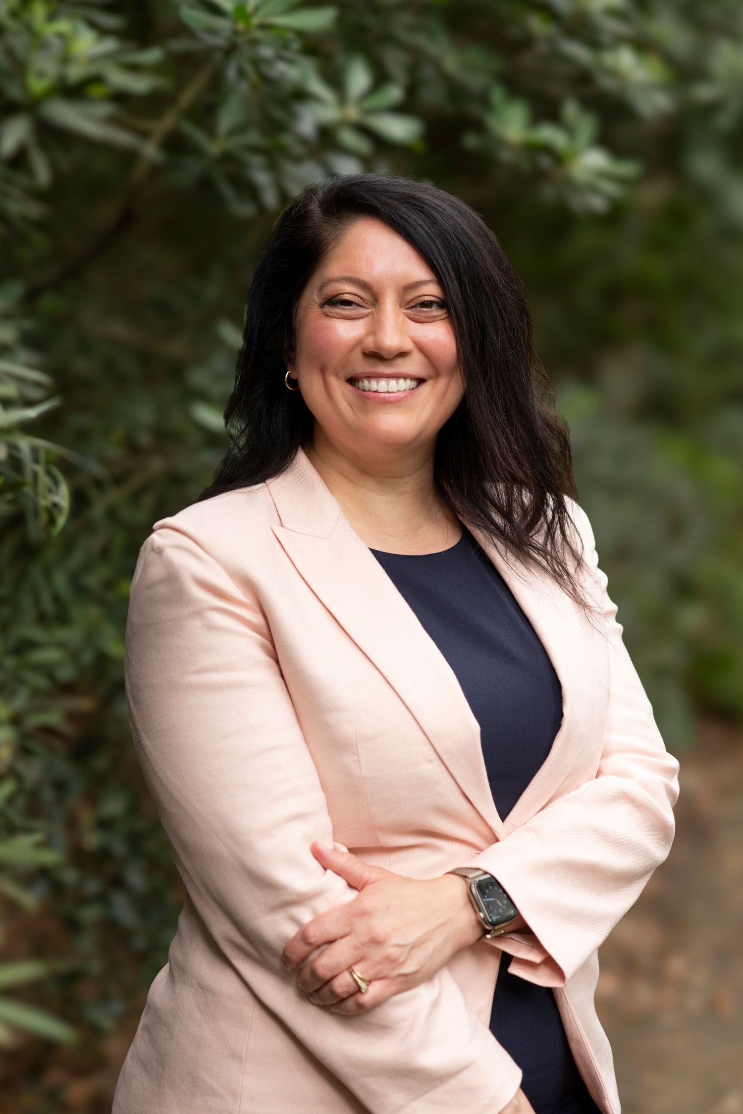 Professional portrait of someone in a light pink blazer and navy top smiling warmly against a green foliage background.