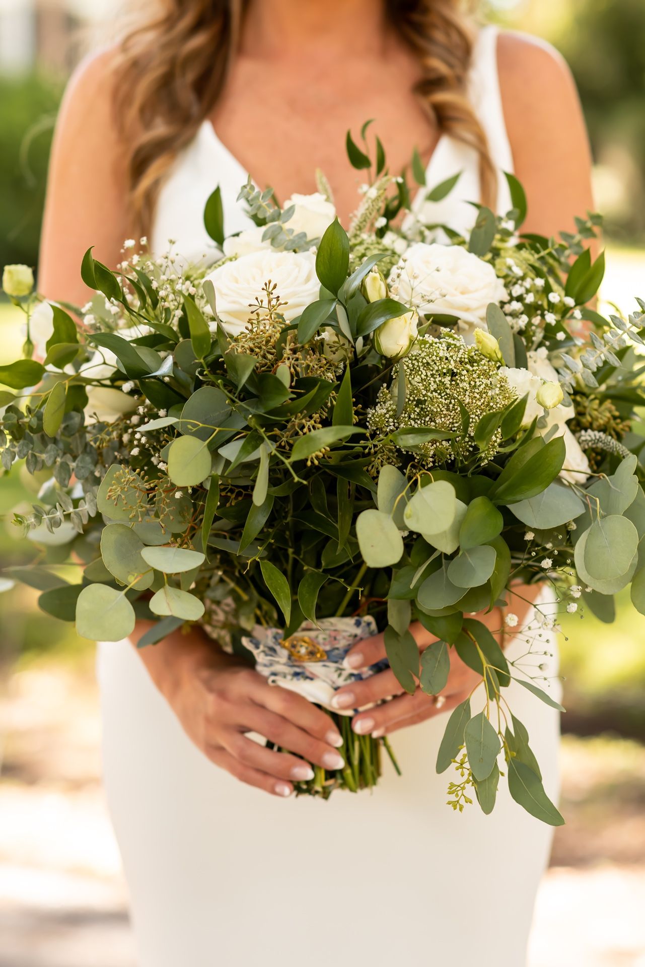 Elegant white and green wedding bouquet featuring eucalyptus leaves and delicate flowers held against a white dress in sunlight.