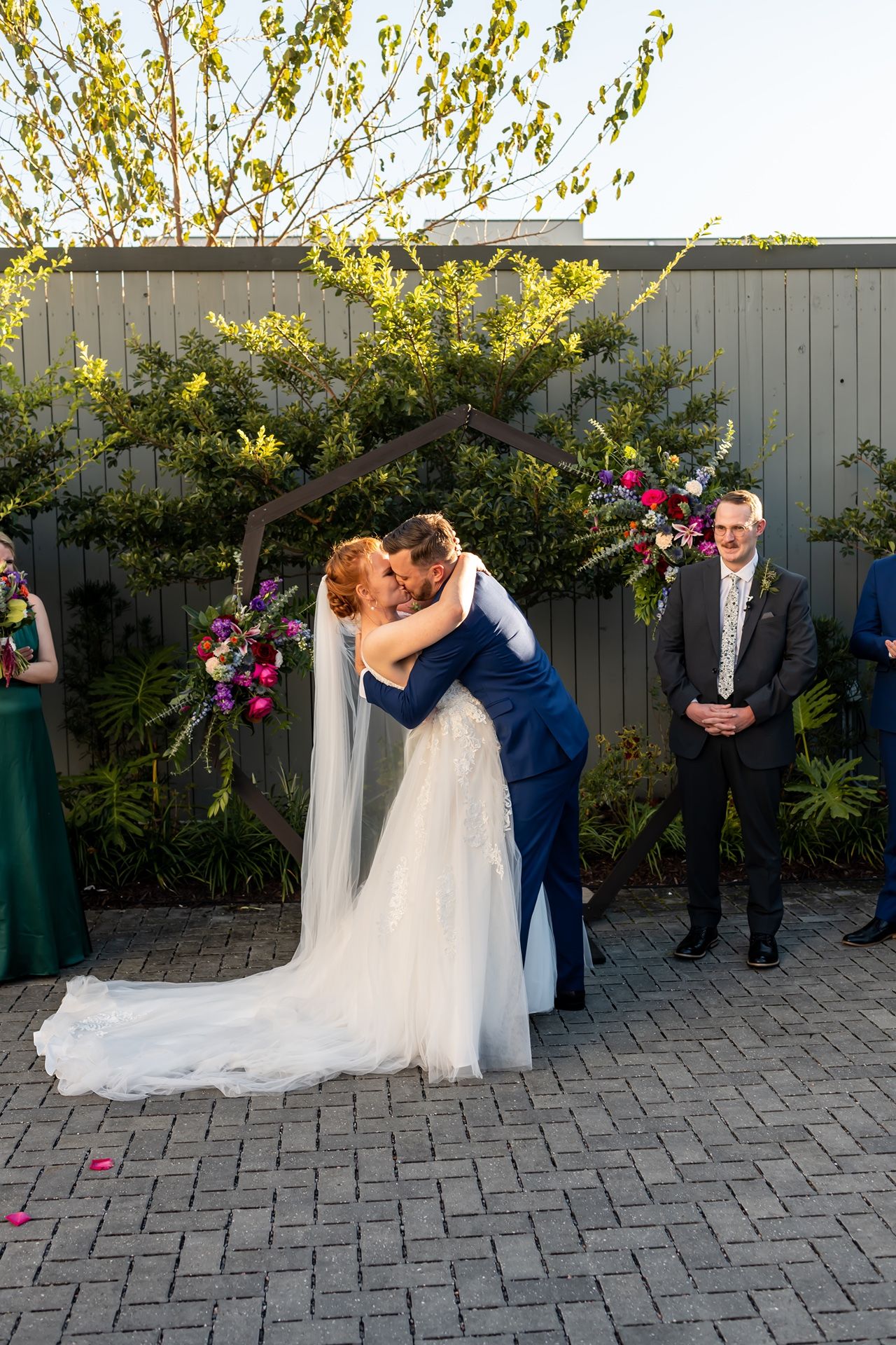 A couple shares a joyful embrace at their outdoor wedding ceremony with flowering trees and guests looking on.