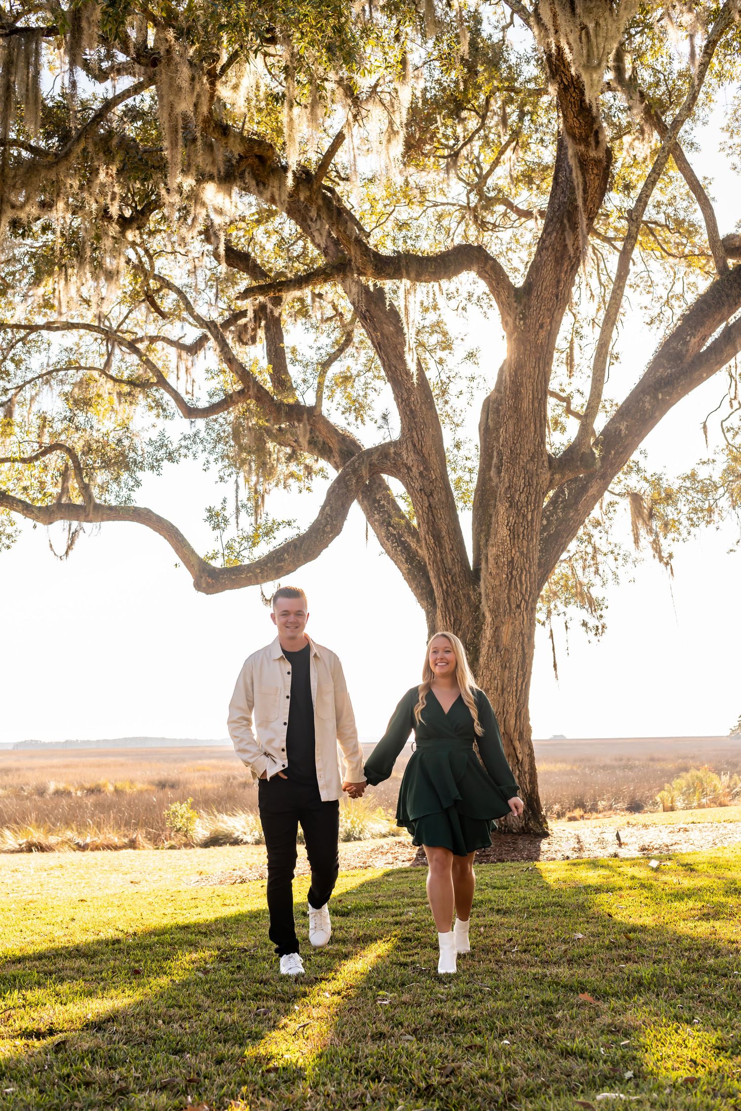 Couple holding hands while walking under large oak tree with Spanish moss at sunset on grassy hilltop.