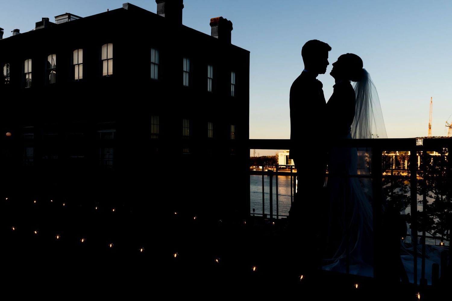 A silhouetted couple stands against a waterfront cityscape at sunset with a dark building in the background.