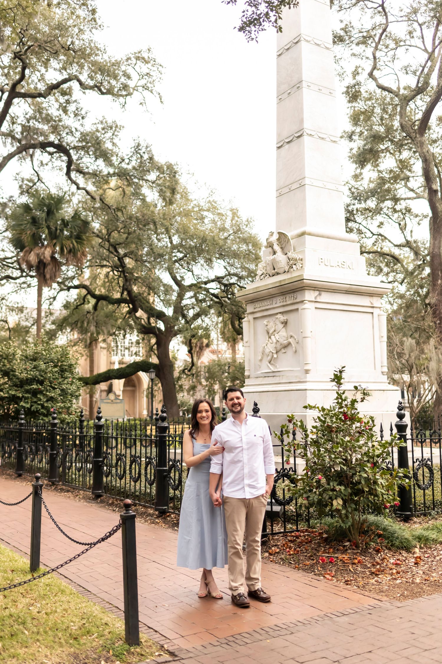 Two people standing by white stone monuments with wrought iron fencing along a brick pathway under Spanish moss-draped trees.