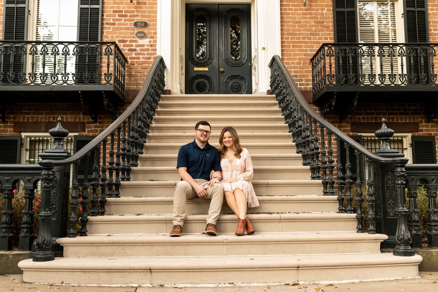 A couple sits together on limestone stairs leading to a historic brick townhouse with ornate black iron railings.