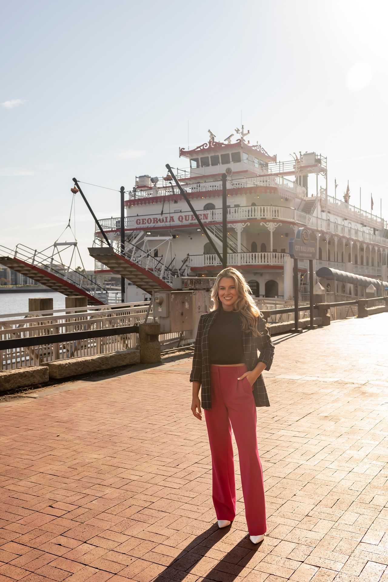 Sunlit tourist standing on red brick boardwalk along historic Natchez riverfront with classic steamboat in background.