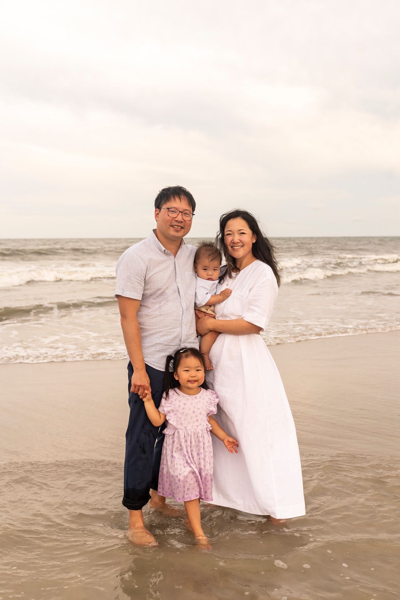 A family stands together at the beach during sunset, barefoot in the sand as waves crash behind them in soft lighting.