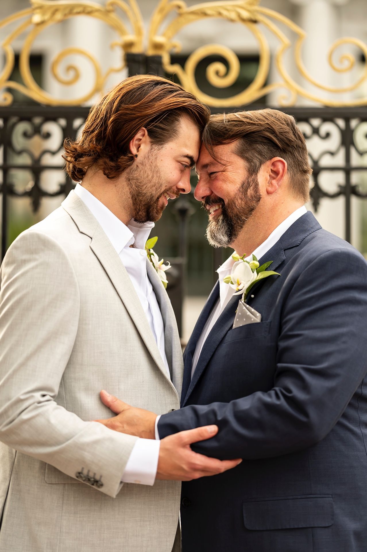 Two people in suits share an intimate moment in front of an ornate iron gate wearing matching boutonnieres.