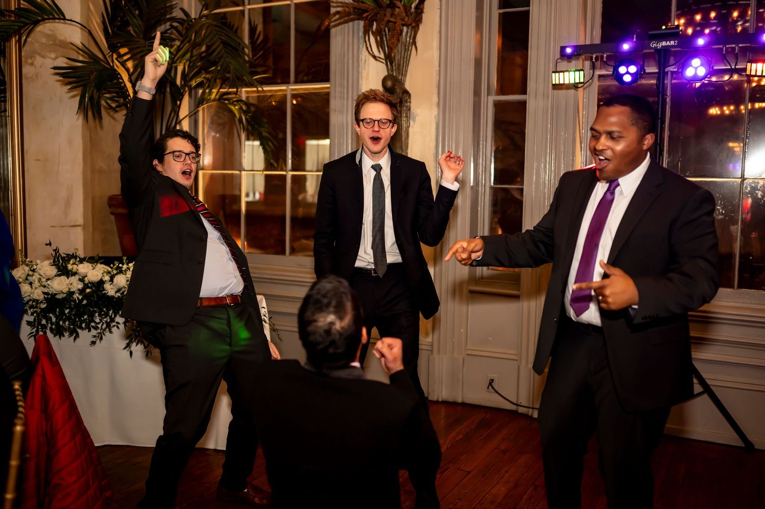 Groomsmen dancing together at a wedding reception in an elegant venue with decorative lights.