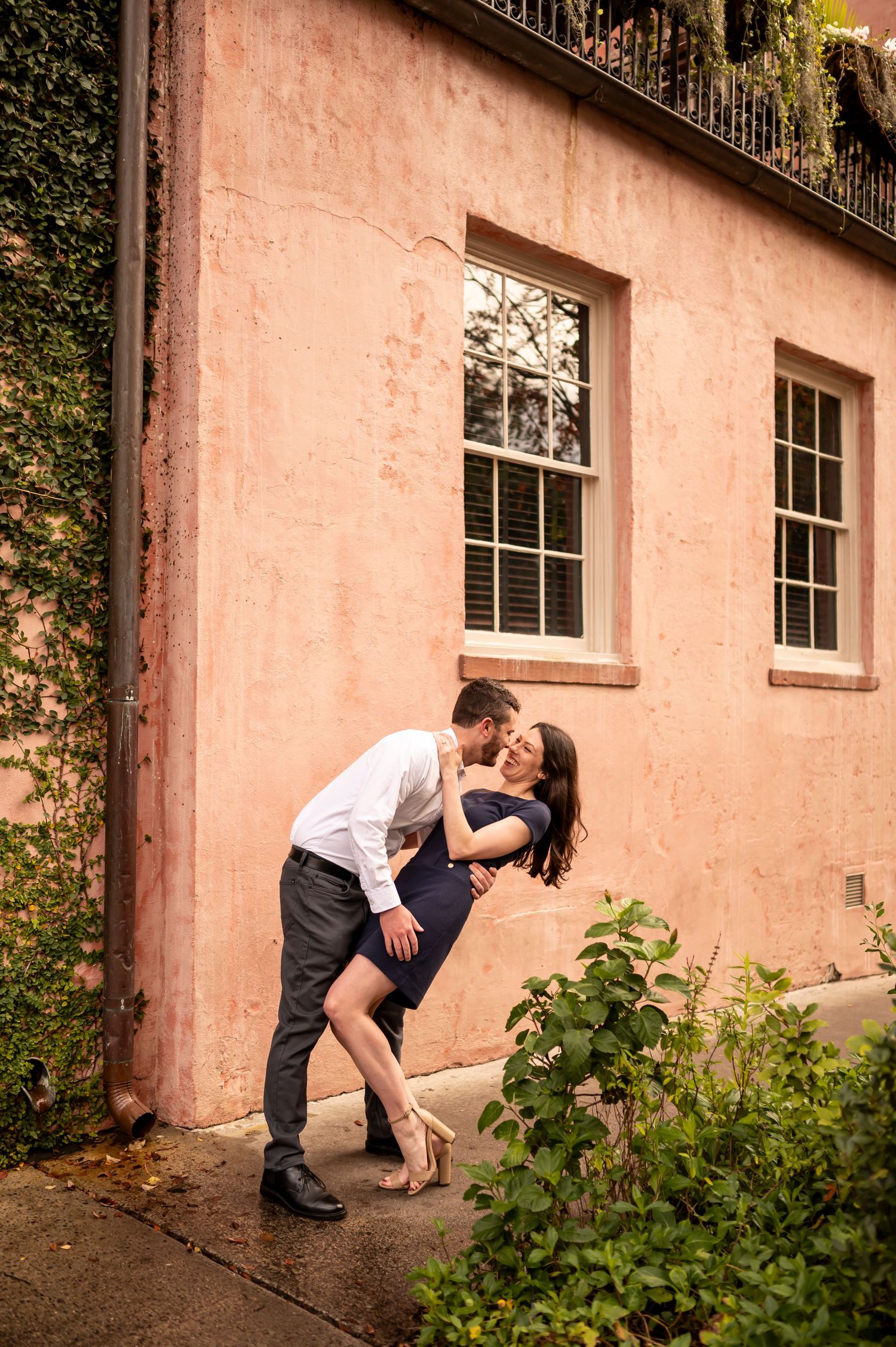 A romantic couple in elegant attire share an intimate moment against a pink stucco wall with climbing vines.