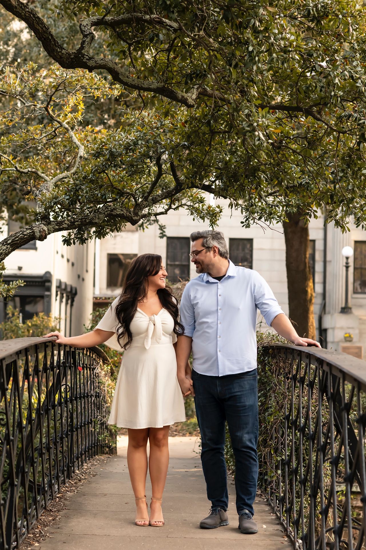 A couple stands together on a charming footbridge surrounded by overhanging tree branches in an urban setting.