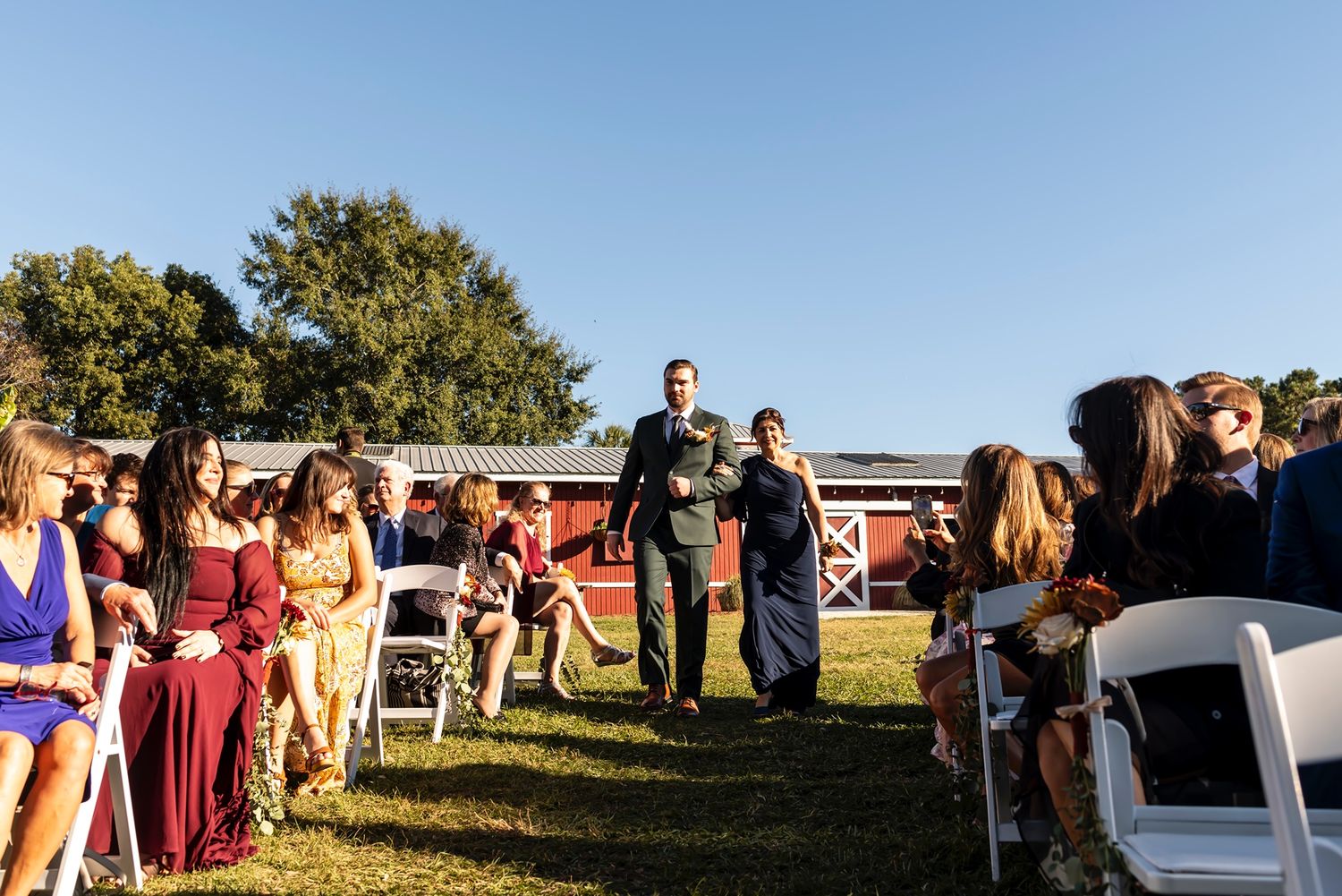 Wedding guests are seated on white chairs outdoors for a ceremony under blue skies and autumn trees.