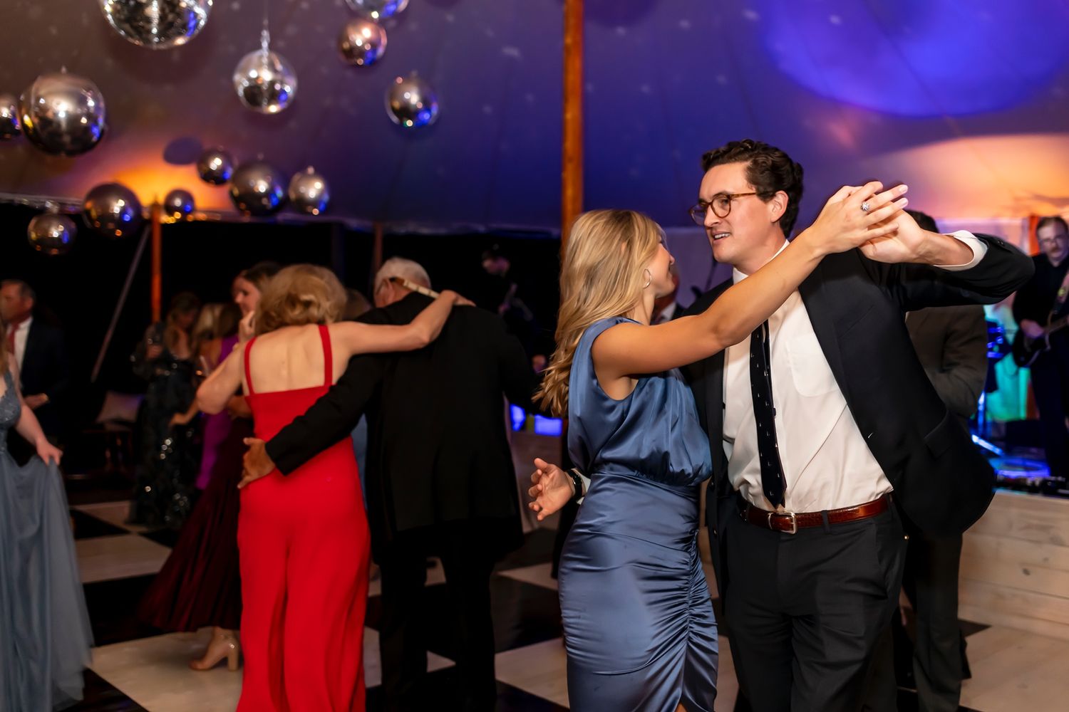 People dance together on a dimly lit dance floor under colorful blue and purple lighting at a social event.