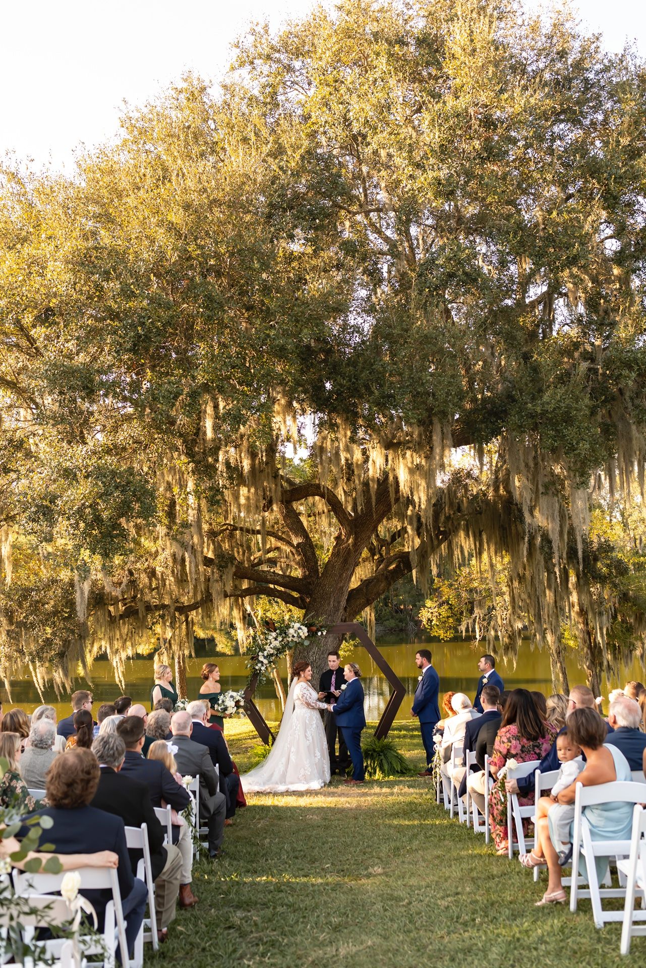 Outdoor wedding ceremony under a majestic Spanish moss-draped oak tree with guests seated on white chairs.