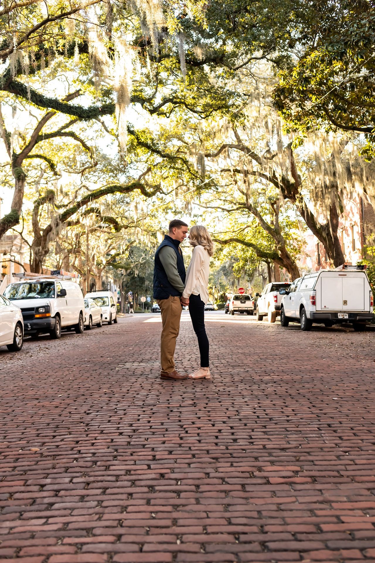 A couple stands on a picturesque brick street lined with Spanish moss-draped oak trees and parked cars in Savannah.