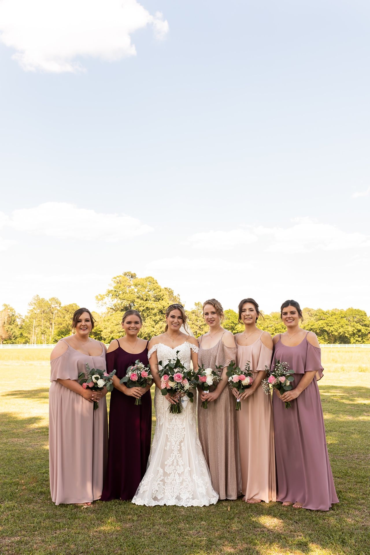 A bridal party poses on grass wearing elegant gowns in shades of burgundy, pink and mauve while holding floral bouquets.