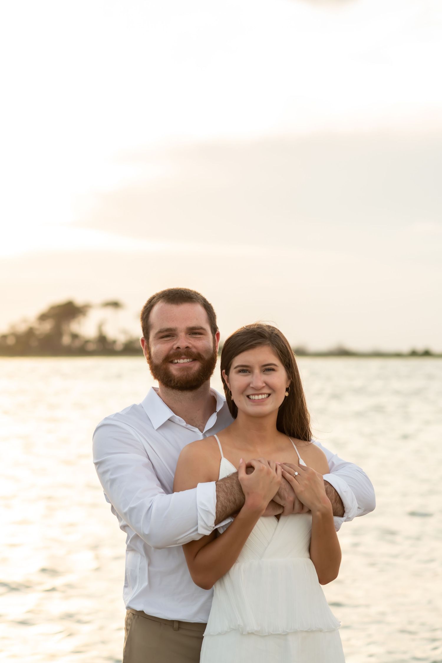 A couple embraces on a beach at sunset with golden light reflecting off the water in the background.